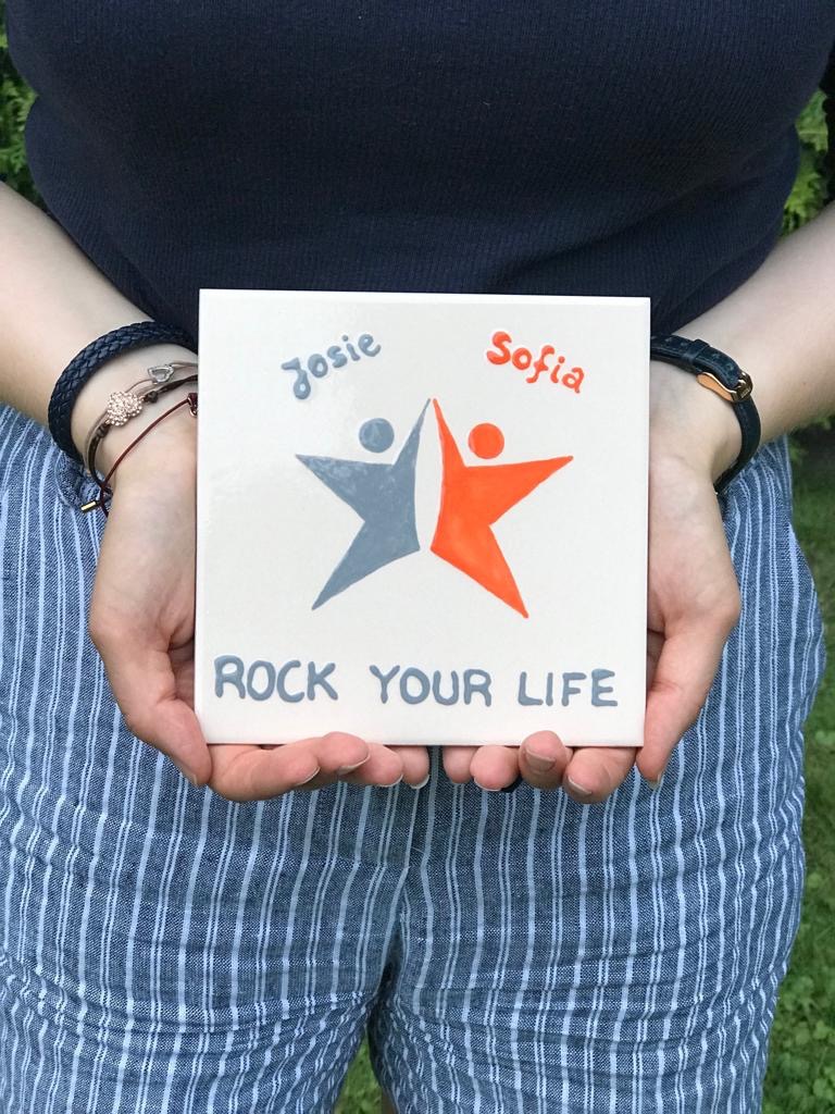 Hände halten eine Fliese, die mit zwei stilisierten Figuren bemalt ist, über denen "Josie" und "Sofia" steht. Darunter steht "Rock your life"