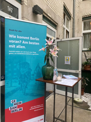 Ein Tisch mit einer Blumenvase vor einer geöffneten Tür, daneben ein Rollup mit dem Text "Stadt für alle: Wie kommt Berlin voran? Am besten mit allen." und Logo GEMEINSAM BERLIN