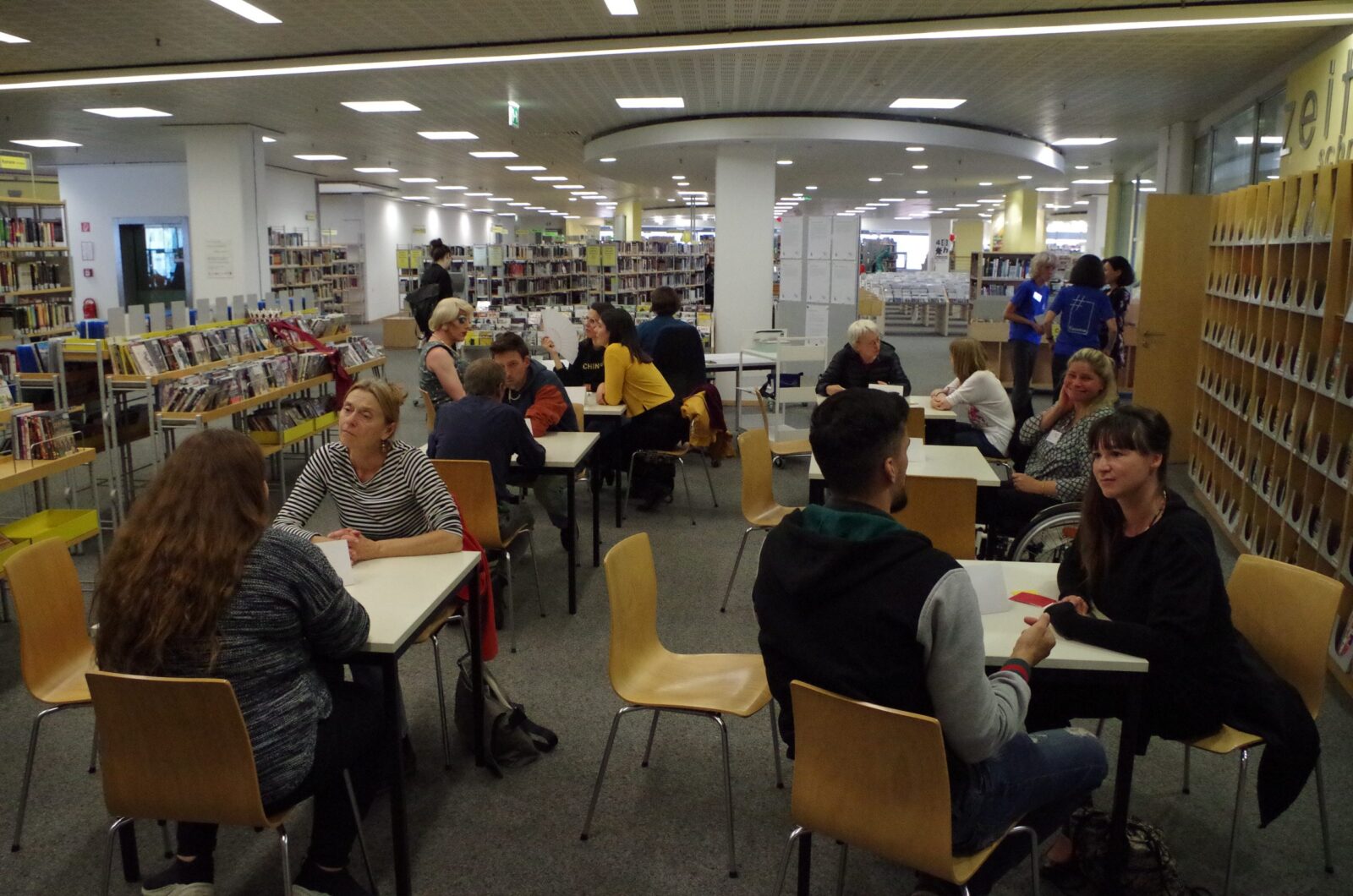Einige Personen unterhalten sich zu zweit an Tischen in einer Bibliothek.